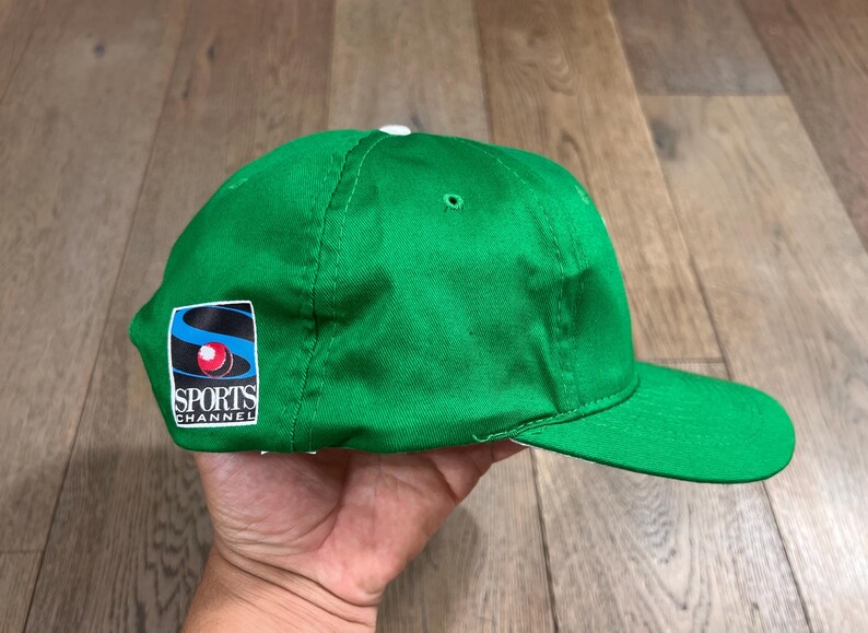 May include: A bright green baseball cap with a curved brim. The cap features a patch on the side with the text "SPORTS CHANNEL" and a logo with blue, red, and white elements. The cap is made of a smooth fabric.