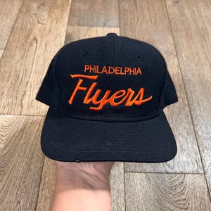 May include: A black baseball cap featuring the text PHILADELPHIA Flyers embroidered in orange thread on the front crown. The cap has a structured six-panel design with a curved brim and eyelets for ventilation on the top panels.