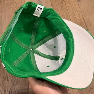 May include: Green baseball cap with a white brim and interior. The cap's interior features a white fabric with green stitching and a small tag. The cap is made of a durable material and is designed for everyday wear.