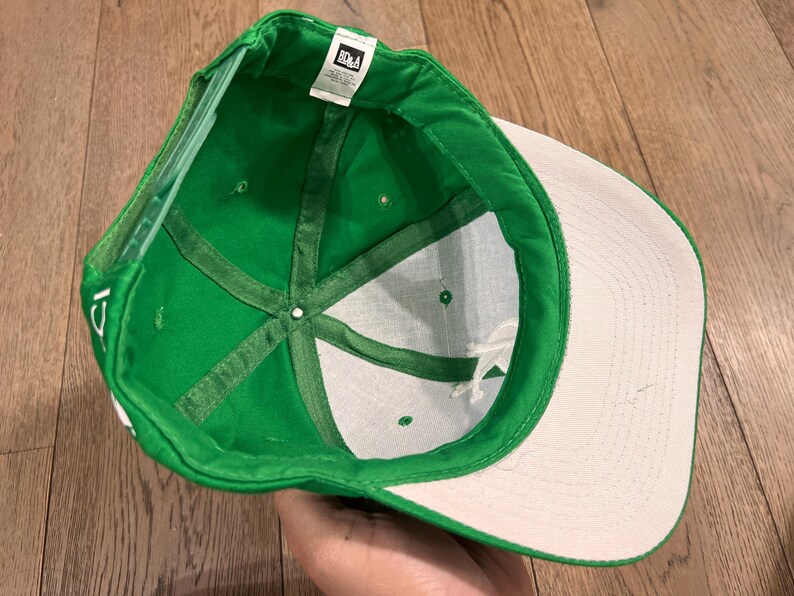 May include: Green baseball cap with a white brim and interior. The cap's interior features a white fabric with green stitching and a small tag. The cap is made of a durable material and is designed for everyday wear.