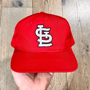May include: A red baseball cap featuring a white and black logo. The cap has a curved peak and a structured crown. The logo is a stylized "STL" design. The cap is made of a durable material and is likely for sports fans.