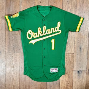 May include: Green baseball jersey with the word "Oakland" in white script across the chest, with a yellow outline. The number "1" is on the front. Yellow and white stripes accent the sleeves. The jersey is button-down.