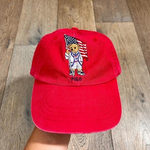 May include: A bright red baseball cap featuring an embroidered graphic of a teddy bear wearing a white blazer and shorts while holding an American flag. The word POLO is stitched in navy blue thread directly below the bear on the front panel.