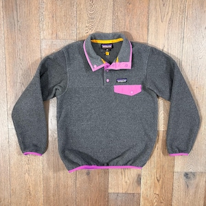 May include: A gray fleece pullover with a pink pocket and collar trim. The collar has a yellow interior and pink buttons. The cuffs and hem are trimmed in pink. The Patagonia logo is visible on the pocket.