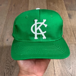 May include: A green baseball cap with a white "KC" logo. The cap has a flat brim and a white button on top. The hat is made of a smooth fabric and is displayed on a wooden surface.