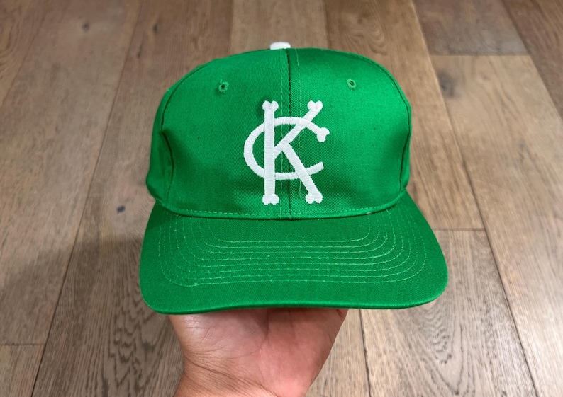 May include: A green baseball cap with a white "KC" logo. The cap has a flat brim and a white button on top. The hat is made of a smooth fabric and is displayed on a wooden surface.