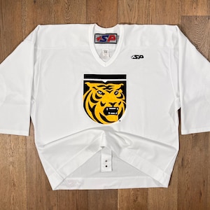 May include: White hockey jersey with a V-neck and long sleeves. The jersey features a black and yellow tiger head logo on the front. The jersey has the letters "SP" on the collar and a small logo on the right chest.