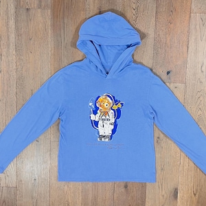 Puede incluir: Una camiseta de manga larga con capucha azul claro con un gráfico de un oso con un traje de buceo. El oso lleva un casco de buceo amarillo y sostiene una lanza. La camiseta tiene el texto "POLO BEAR by RALPH LAUREN" y una firma.