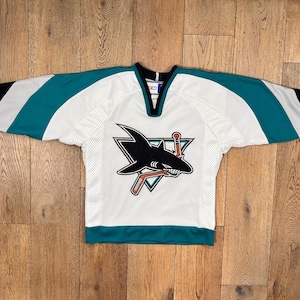 Puede incluir: Camiseta de hockey blanca con detalles turquesa y negros. La camiseta presenta un logotipo de tiburón negro con un palo de hockey y un triángulo. Las mangas tienen rayas turquesas y puños negros. El cuello es negro.