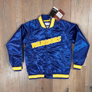 May include: A blue satin jacket with the word "WARRIORS" in yellow block letters across the chest. The jacket has yellow and white striped trim on the collar, cuffs, and waist. A tag is attached.