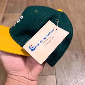 May include: A dark green baseball cap with a bright yellow brim and a yellow button on top. A white paper tag is attached to the side, featuring the blue and red Sports Specialties logo and the text P.O. BOX 16565 IRVINE, CALIF. 92713.