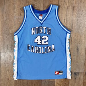 May include: Light blue basketball jersey with the words "NORTH CAROLINA" and the number "42" in white. The jersey has a V-neck and white and blue trim. A Nike logo is visible on the chest and a tag at the bottom.