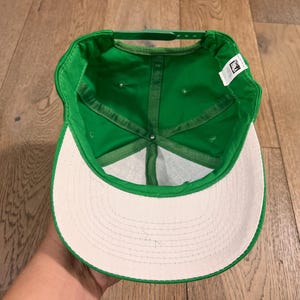 May include: A green baseball cap with a white brim, viewed from the inside. The cap has a structured crown and a curved brim. The interior features a green sweatband and stitching. A small tag is visible.