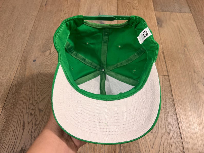 May include: A green baseball cap with a white brim, viewed from the inside. The cap has a structured crown and a curved brim. The interior features a green sweatband and stitching. A small tag is visible.