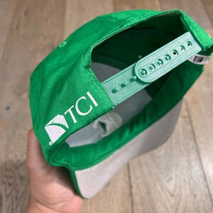 May include: Green baseball cap with a gray brim and adjustable strap. The cap features the white text "TCI" on the side. The cap is made of a durable fabric and has a classic snapback closure.
