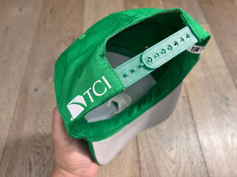 May include: Green baseball cap with a gray brim and adjustable strap. The cap features the white text "TCI" on the side. The cap is made of a durable fabric and has a classic snapback closure.