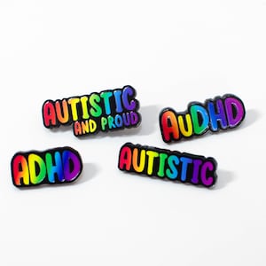 May include: Four enamel pins with black borders and rainbow gradient lettering. The pins feature the text "AUTISTIC AND PROUD", "AuDHD", "ADHD", and "AUTISTIC" in a bold, rounded font style, showcasing vibrant colours against a plain white background.