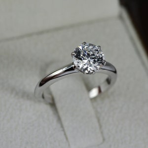 May include: A silver engagement ring with a round, clear diamond in a white velvet ring box.