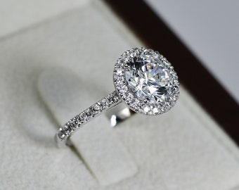 May include: A silver engagement ring with a large, round, clear center stone surrounded by a halo of smaller diamonds. The ring is set in a white gold band and is displayed in a brown velvet ring box.