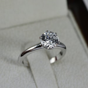 May include: A silver engagement ring with a round, clear diamond in a white velvet ring box.