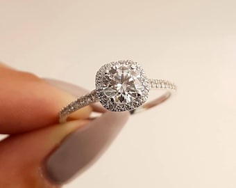 Op de afbeelding: Een zilveren verlovingsring met een ronde, heldere middelste steen omgeven door een halo van kleinere diamanten. De band is ook bezet met kleine diamanten.