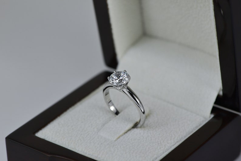 May include: A silver engagement ring with a large, round, clear diamond in a black ring box.