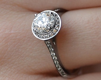 Op de afbeelding: Een zilveren verlovingsring met een grote, ronde, heldere diamant in het midden, omgeven door een halo van kleinere diamanten. De band is ook bezet met kleinere diamanten.