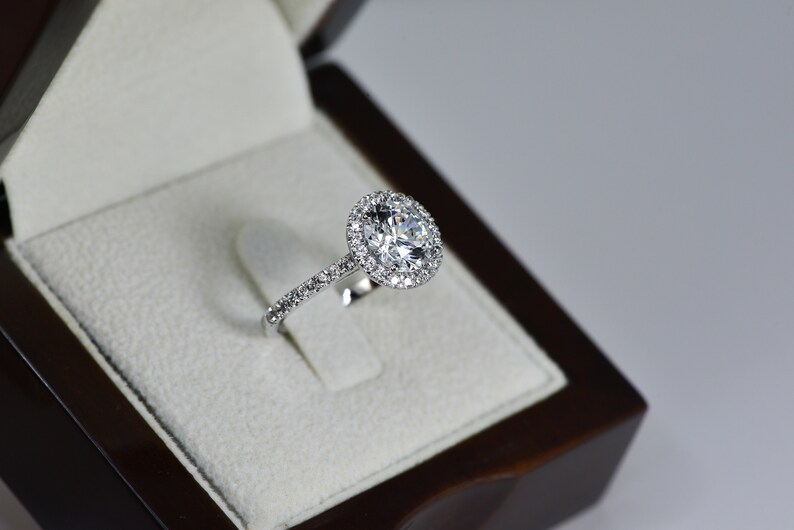K&ouml;nnte beinhalten: Ein silberner Verlobungsring mit einem gro&szlig;en, runden, klaren Diamanten, der von einem Heiligenschein aus kleineren Diamanten umgeben ist. Der Ring befindet sich in einer wei&szlig;en Samtringschatulle.