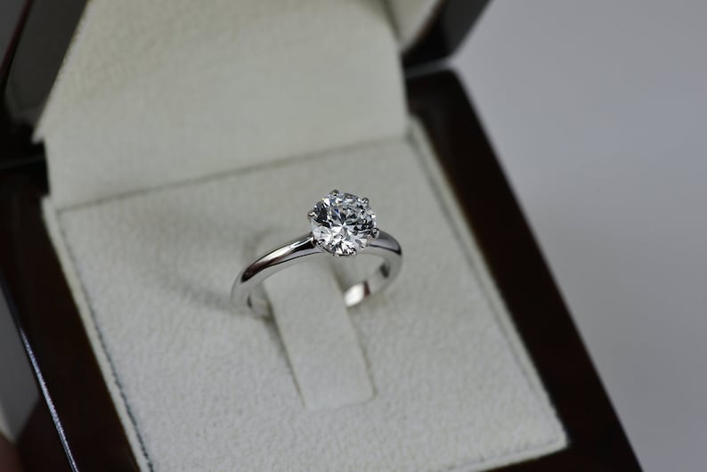 May include: A silver engagement ring with a round, clear diamond in a white velvet ring box.