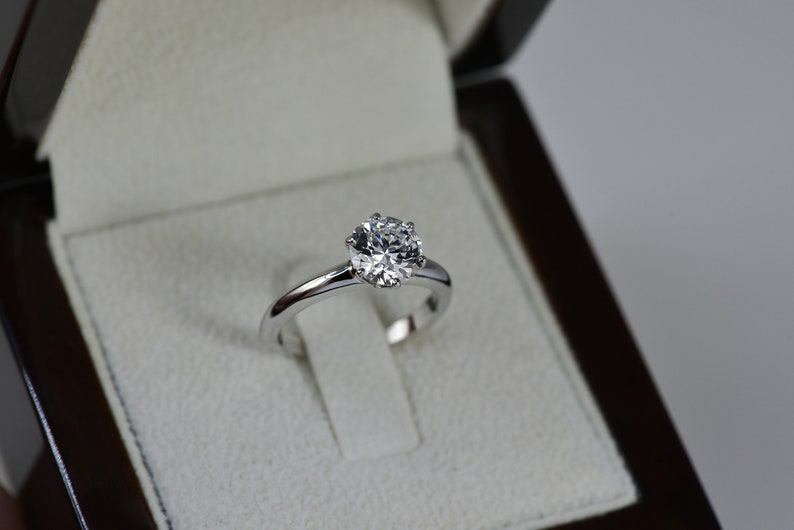 May include: A silver engagement ring with a round, clear diamond in a white velvet ring box.