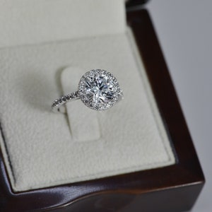 K&ouml;nnte beinhalten: Ein silberner Verlobungsring mit einem gro&szlig;en runden Diamanten in der Mitte, umgeben von einem Heiligenschein aus kleineren Diamanten. Der Ring befindet sich in einer wei&szlig;en Samt-Ringbox.