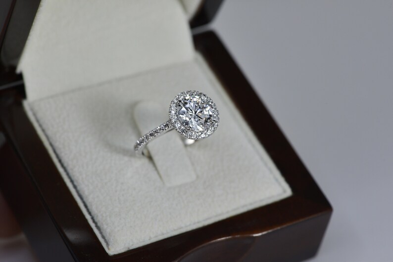 K&ouml;nnte beinhalten: Ein silberner Verlobungsring mit einem gro&szlig;en runden Diamanten, der von einem Heiligenschein aus kleineren Diamanten umgeben ist. Der Ring befindet sich in einer wei&szlig;en Samtringbox.