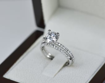 May include: A silver engagement ring set with a large, round, clear center stone and a double band of smaller, clear stones.