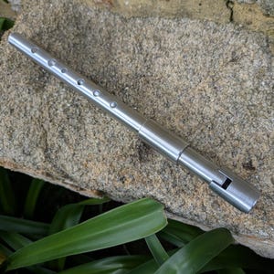 Può includere: Un flauto in metallo color argento con diversi piccoli fori lungo la sua lunghezza. Il flauto è appoggiato su una superficie rocciosa grigia e strutturata, con fogliame verde in primo piano.