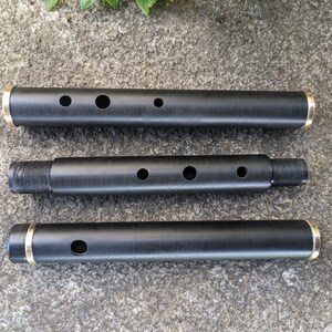 May include: Three black wooden flute sections with gold-colored metal rings at the ends. The sections are laid out horizontally on a gray textured surface.
