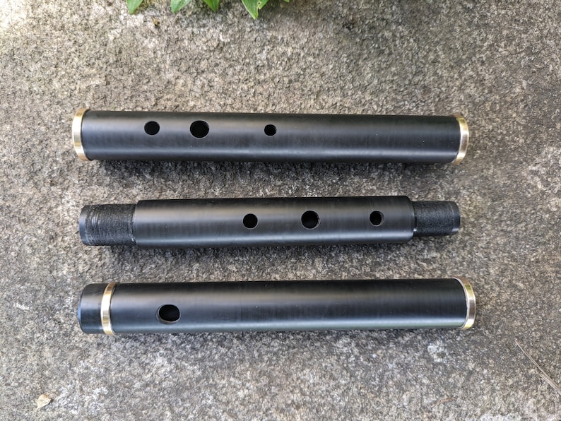 May include: Three black wooden flute sections with gold-colored metal rings at the ends. The sections are laid out horizontally on a gray textured surface.