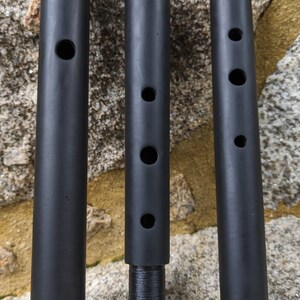 May include: Three black cylindrical tubes with different numbers of holes drilled into them. The tubes are made of a smooth, black material and are arranged in a row from left to right.