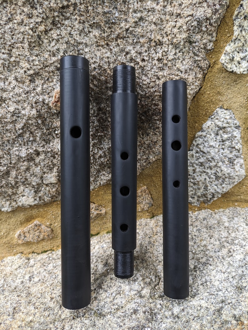 May include: Three black metal rods with holes drilled into them. The rods are arranged vertically against a stone background.