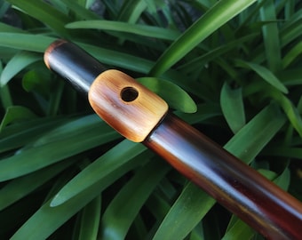 May include: A wooden flute with a dark brown stain and a natural wood mouthpiece. The flute is lying on a bed of green leaves.