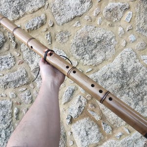 Puede incluir: Flauta de bambú con acabado de madera natural, con múltiples agujeros para los dedos y bandas negras decorativas en los extremos. El instrumento se sostiene contra un fondo de pared de piedra.