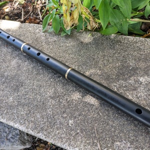 May include: A black wooden Irish flute with gold accents. The flute has a series of holes along the top and a mouthpiece at one end.