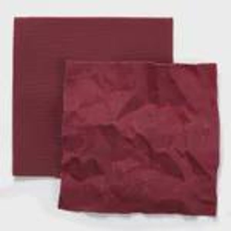 Op de afbeelding: Twee vierkante vellen donkerbordeauxrood papier. Het ene vel ligt achter het andere en ziet er glad en plat uit. Het voorste vel is verfrommeld, waardoor een getextureerd oppervlak met diverse vouwen en kreukels over het hele vierkante vlak ontstaat.