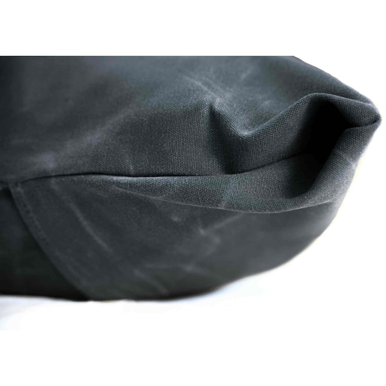Op de afbeelding: Een donker antracietgrijze canvas tas met een dikke, duurzame textuur en zichtbare vouwen en kreukels. Het materiaal heeft een matte afwerking en een stevige constructie, waarbij een hoekdetail van de tas met stiksels zichtbaar is.