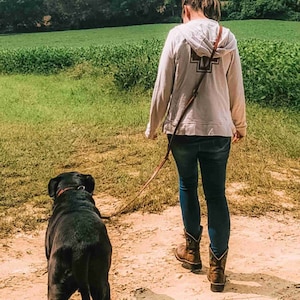 May include: A person walks with a black dog on a leash in a field. The person is wearing a white hoodie with a brown leather strap and blue jeans. The dog is walking in front of the person.
