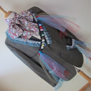 May include: A brown plaid blazer with a white and blue patterned scarf, a lace trim, and a colorful string of spools of thread. The blazer is decorated with blue, pink, and white tulle.