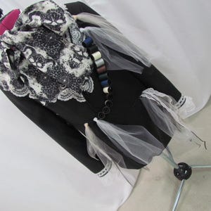 May include: A black jacket with white lace trim and a decorative scarf featuring a black and white pattern. The jacket is adorned with small spools of thread and white tulle accents, displayed on a mannequin.