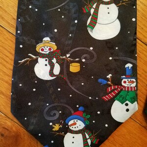 May include: A black necktie with a repeating pattern of snowmen holding buckets and birds. The snowmen are wearing colorful scarves and hats. The background is a dark blue with white dots.