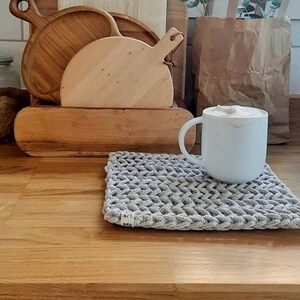 May include: A white ceramic mug with a beige knitted coaster on a wooden surface. The mug is filled with a light brown beverage. There are several wooden chopping boards stacked in the background.
