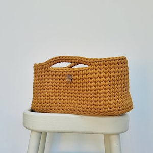 May include: A mustard yellow crocheted storage basket with two handles. The basket is sitting on a white wooden stool.
