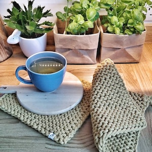 May include: Two green knitted pot holders with a white tag that says "Knit Art" on each. The pot holders are on a wooden surface with a blue mug of tea and a wooden chopping board.